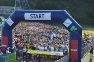 Startlinie des Firmenlaufs Thüringens Süden, mit einer großen Menschenmenge von Läufern in bunten Laufshirts, die gespannt auf den Beginn des Rennens warten, unter einem großen aufblasbaren Startbogen mit dem Firmenlauf-Logo und Ballons an einem sonnigen Tag.